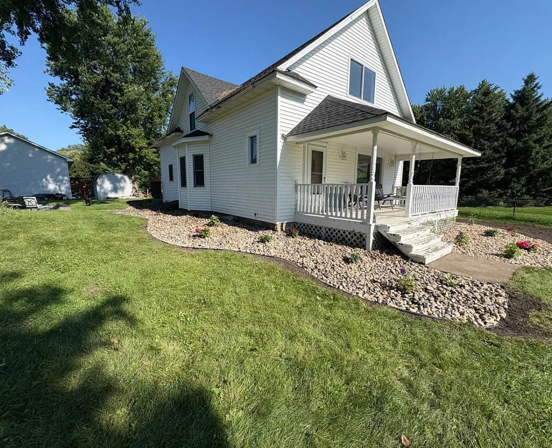 White two-story house with porch, landscaped lawn, and stone border in bright daylight.