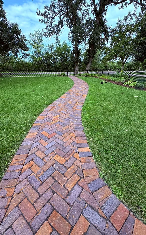 Curved brick pathway through green lawn with trees and fence in the background.